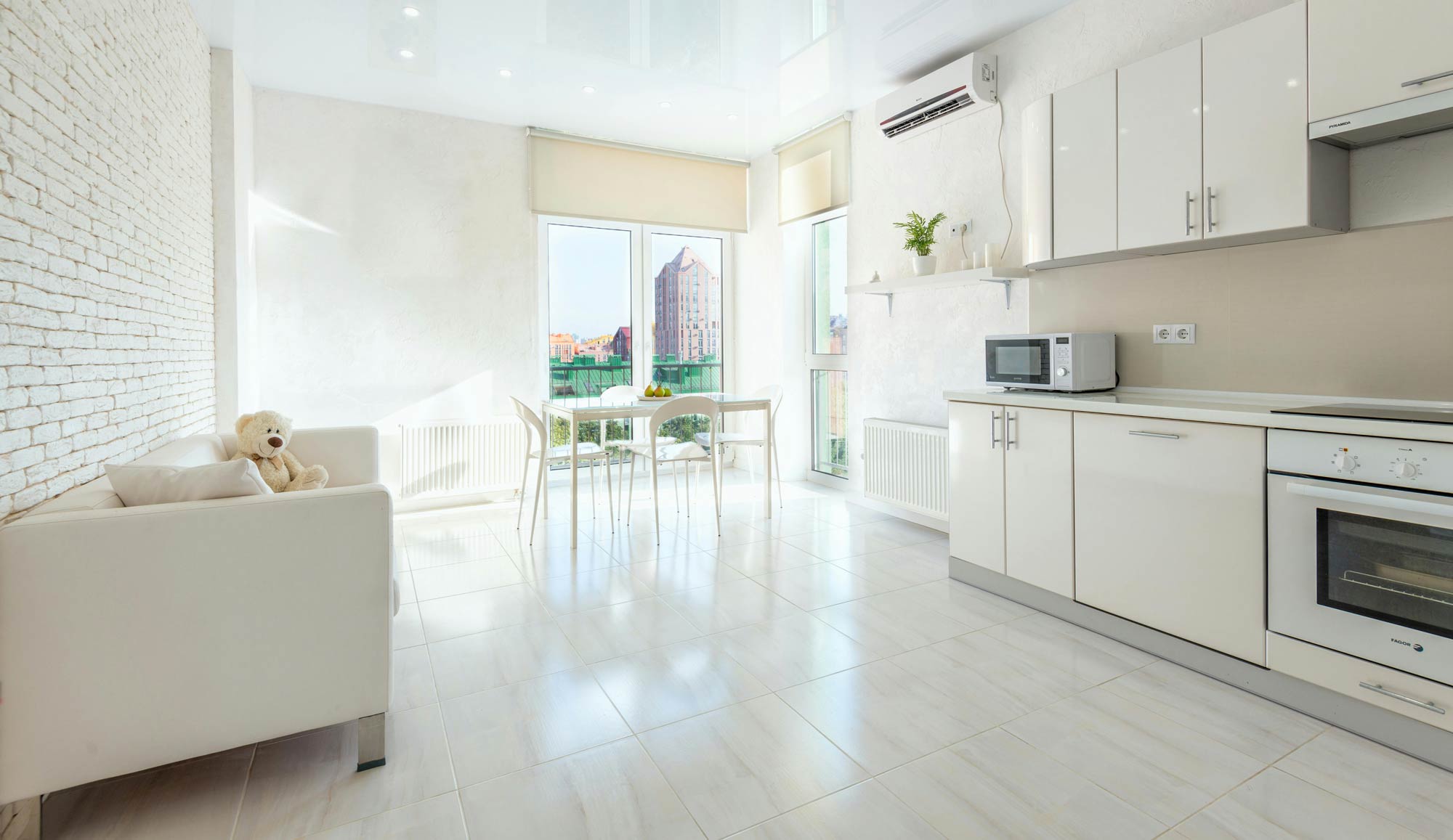 Světlý minimalistický byt s bílým nábytkem, stolem u okna a výhledem na město.