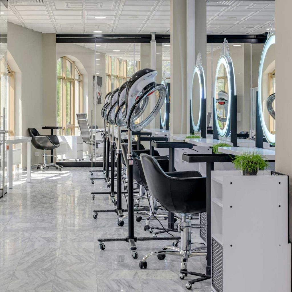 Moderní a světlý kadeřnický salon s řadou křesel, osvětlenými zrcadly a sušáky na vlasy.