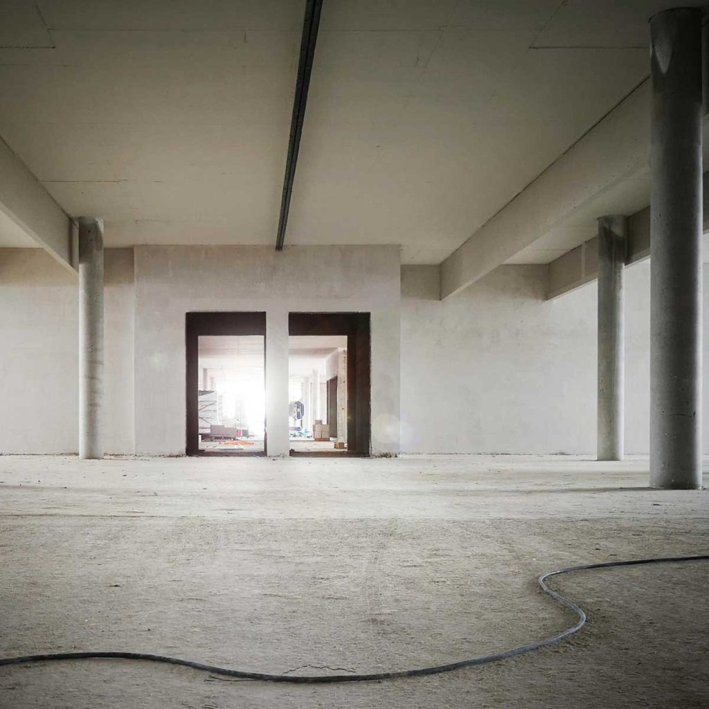 Prázdná hala po stavbě s betonovou podlahou, sloupy a rozvody na zemi.